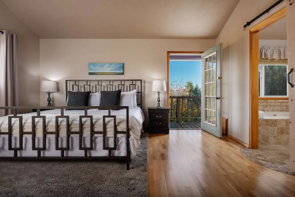 This interior image features a bed with a decorative headboard positioned next to a large window, providing views of the outdoors. There is a door leading directly to a balcony, and the atmosphere is enhanced by serene, natural lighting. The room evokes a peaceful haven, perfect for relaxation, and hints at luxury with the possibility of a private fireplace and a tub nearby.