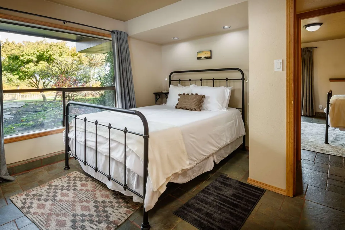 A tranquil bedroom scene with a cozy bed positioned near a large window. The window frames a peaceful view of nature beyond, allowing plenty of natural light to fill the room. Soft textures and calming colors create a restful atmosphere, inviting guests to relax while taking in the scenery outside.