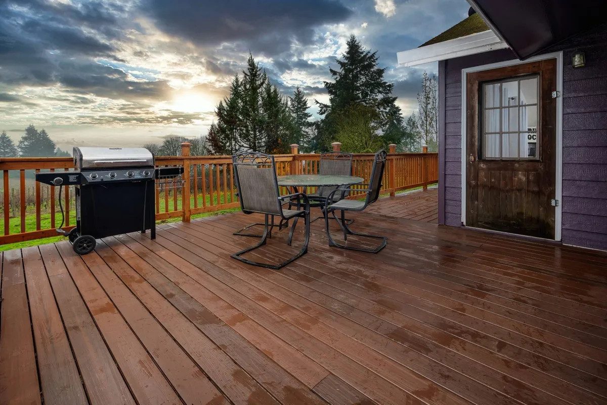 A spacious Fiberon deck set against a backdrop of lush greenery, featuring a premium propane grill and a large outdoor dining table. The wooden railing frames the view, while the setting sun casts a warm, inviting glow, enhancing the sense of comfort and festivity. People are seen barbecuing, dining, and relaxing, emphasizing the social, communal aspect of the space.