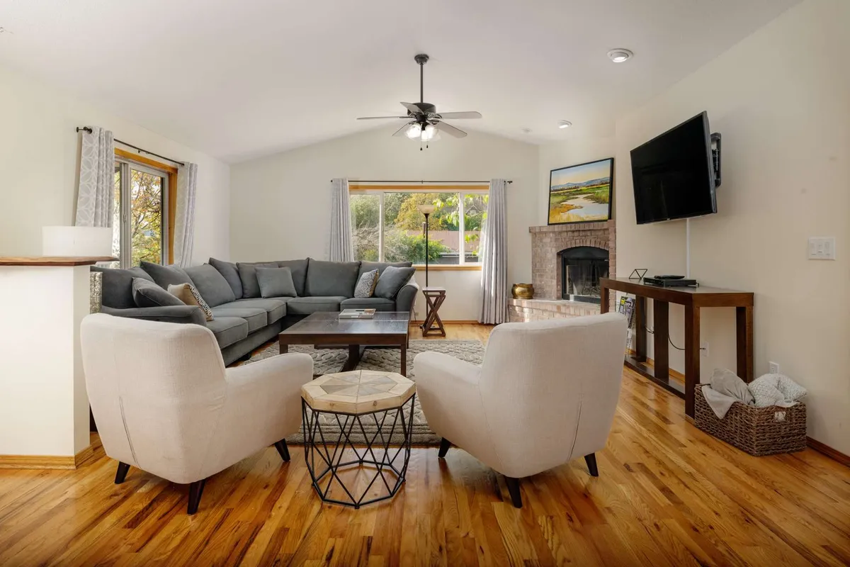 This image showcases a large, open living room with a comfortable sectional sofa arranged around a warm fireplace. Large windows line the wall, offering glimpses of natural light and the scenic surroundings outside. Inviting furnishings and a TV complete the setup, creating a space that is both stylish and conducive to group gatherings.